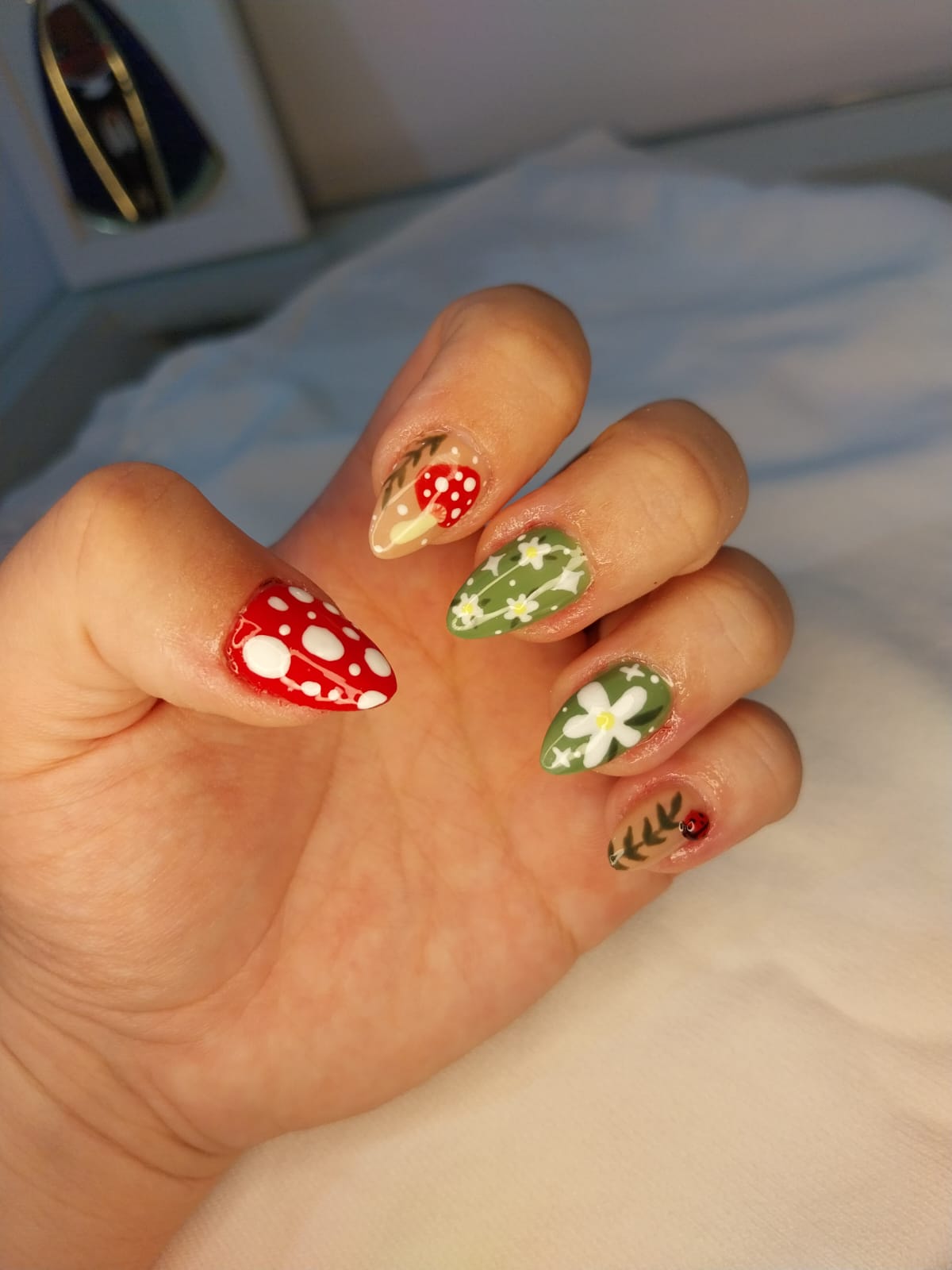 Unhas amendoadas com nail art floral, margaridas, cogumelo e joaninha em tons de verde e vermelho